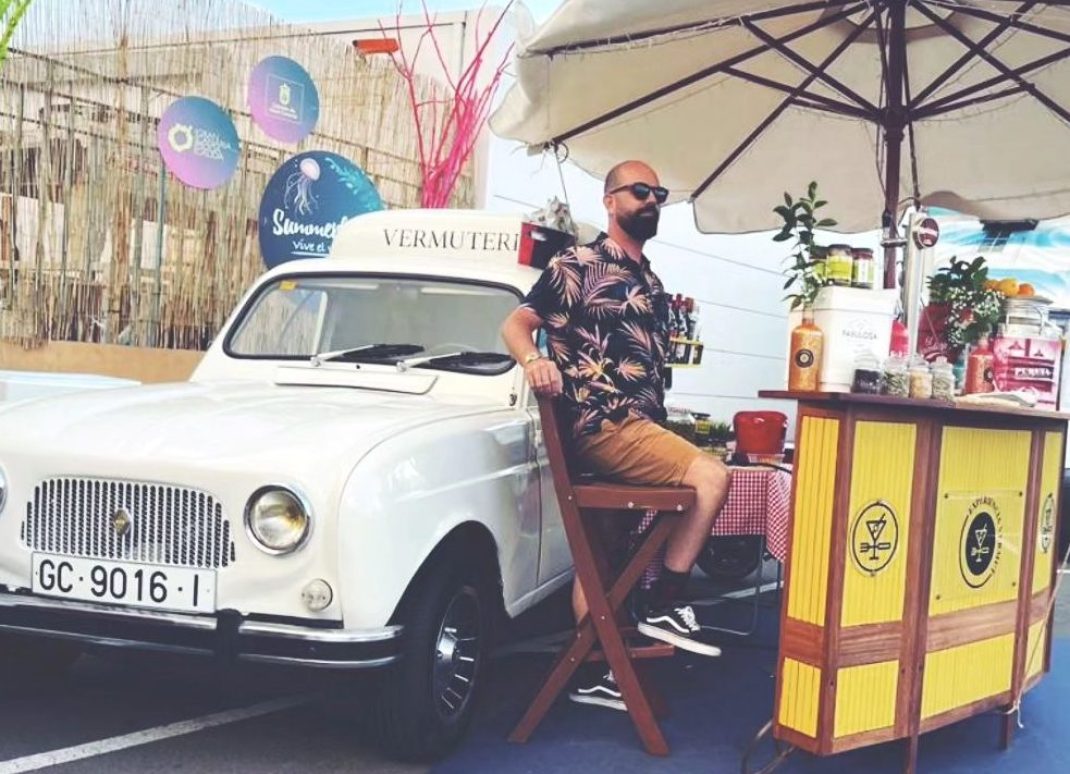 un camarero sentado detras de una barra al aire libre delante de un coche antiguo blanco
